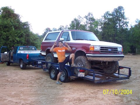 1987 Ford Bronco - A man poses on a trailer carrying a Ford Bronco, with a Chevrolet pickup truck in tow.