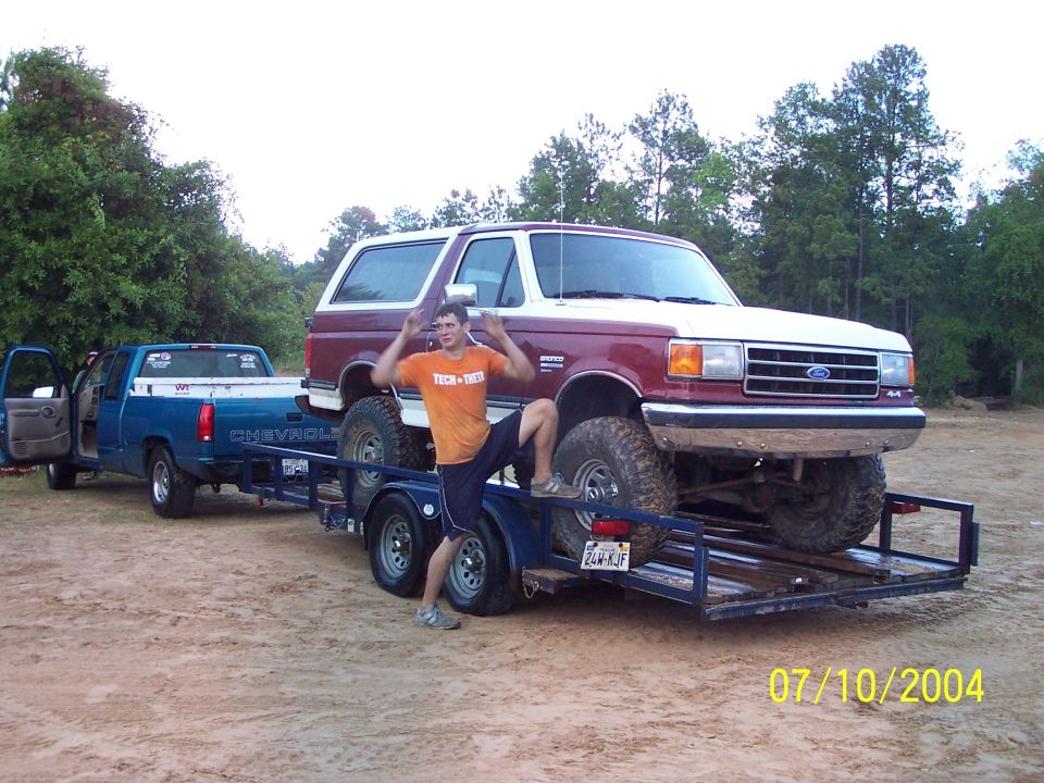 Man posing on trailer carrying a red and white Ford Bronco, blue Chevrolet pickup truck is attached to trailer