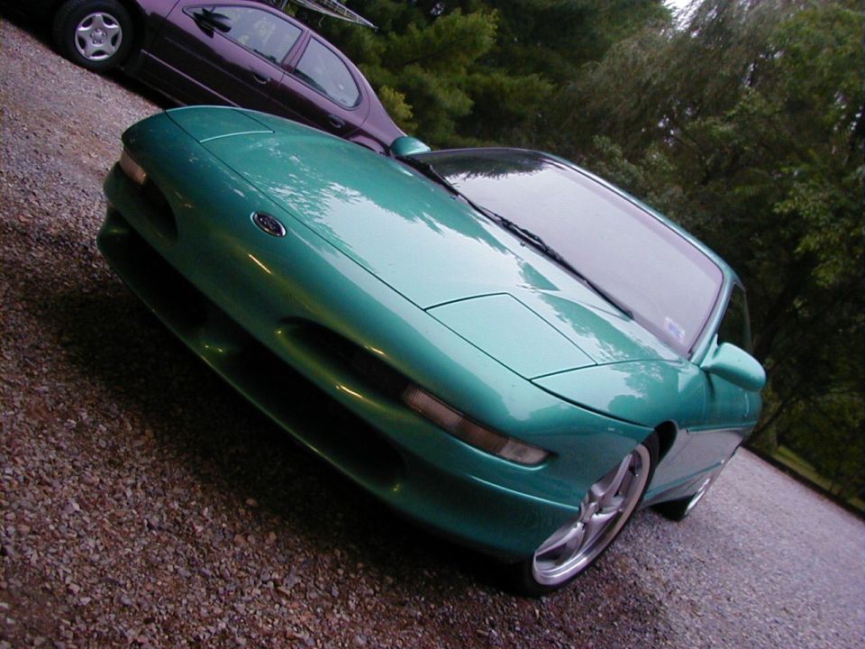 Green coupe with pop-up headlights and aftermarket wheels parked on gravel