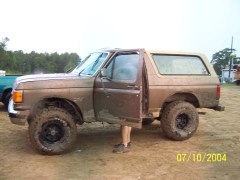 1987 Ford Bronco - A muddy brown 1987 Ford Bronco sits with its driver's door open, showing off its off-road tires.