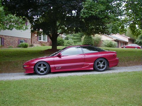 1994 Ford Probe - Side view of a red 1994 Ford Probe GT parked on a gravel driveway in front of houses.