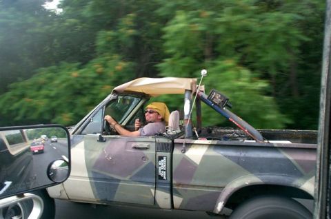 1988 Ford Bronco - A man drives a camouflage pickup truck with a soft top removed on a highway surrounded by trees.