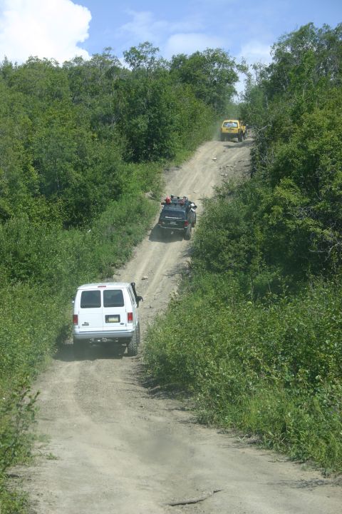 2003 Jeep TJ - Multiple off-road vehicles navigate a challenging dirt path surrounded by dense green foliage under a partly cloudy sky.
