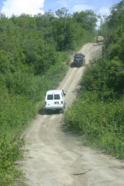 2003 Jeep TJ - Multiple vehicles navigate a steep, dusty dirt road surrounded by dense green foliage.