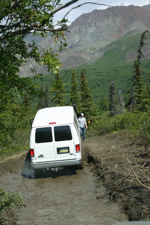 2003 Jeep TJ - A white van struggles on a muddy trail, surrounded by dense forest and mountains.