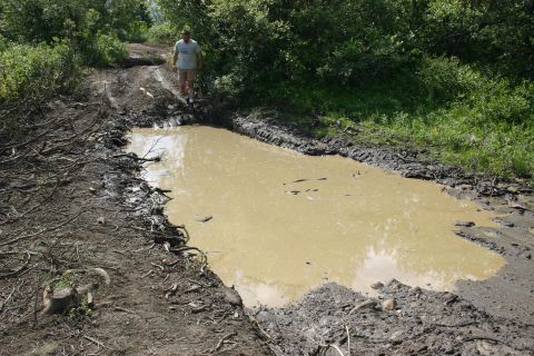 2003 Jeep TJ - A man stands on a muddy, debris-strewn dirt road next to a large, murky puddle.