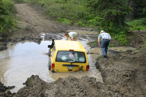 2003 Jeep TJ - Two people attempt to free a yellow 2003 Jeep TJ that is stuck in a muddy water hole on a dirt trail.