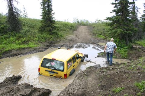 2003 Jeep TJ - A yellow Jeep Cherokee XJ is stuck in a deep mud puddle on an off-road trail, with a man observing the situation.