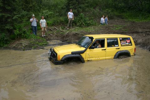 2003 Jeep TJ - A yellow Jeep Cherokee XJ is stuck in a muddy body of water, with onlookers observing from the bank.