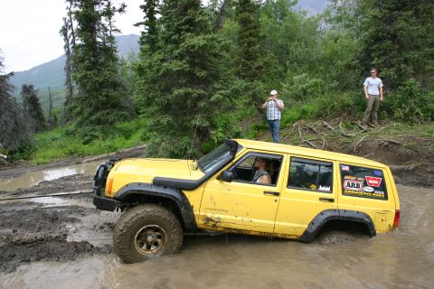 2003 Jeep TJ - Off-roading Jeep Cherokee XJ navigates a deep mud obstacle on a wooded trail.
