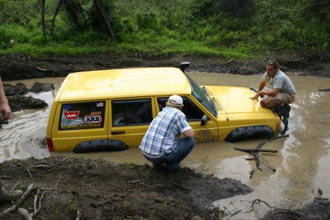 2003 Jeep TJ - Two people attempt to free a yellow Jeep Cherokee that is stuck deep in a muddy puddle.