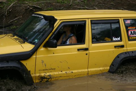 2003 Jeep TJ - A yellow Jeep Cherokee navigates through a muddy off-road course, with a driver visible inside.