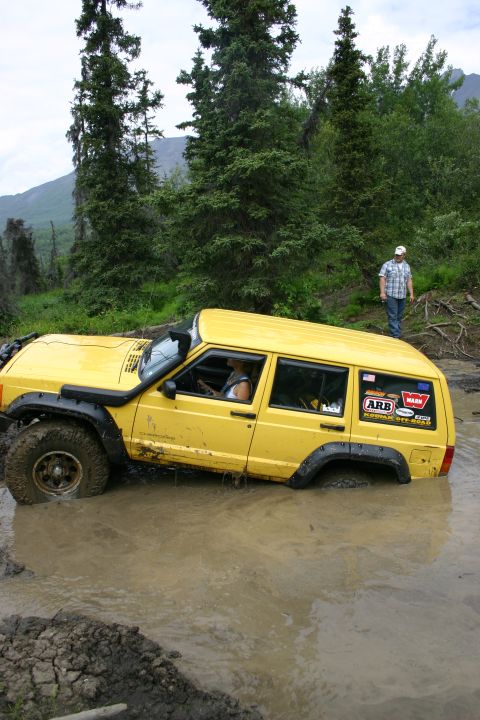 2003 Jeep TJ - A yellow Jeep Cherokee is stuck deep in a muddy puddle, with a person standing on the bank watching.