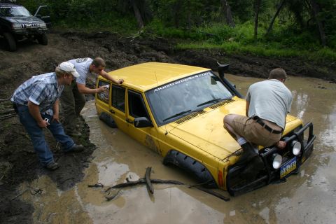 2003 Jeep TJ - Yellow Jeep Cherokee stuck in mud with people trying to help. Another Jeep is in the background.