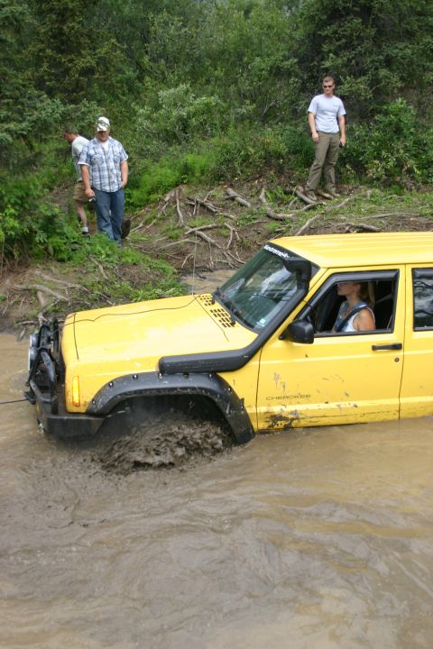 2003 Jeep TJ - A yellow Jeep Cherokee XJ navigates through a muddy water obstacle with onlookers present.