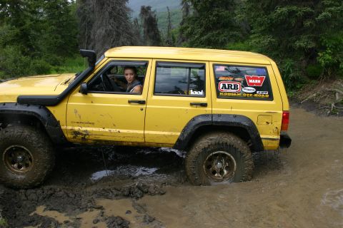 2003 Jeep TJ - Yellow Jeep Cherokee XJ navigates a muddy off-road trail, showing its snorkel and various stickers.