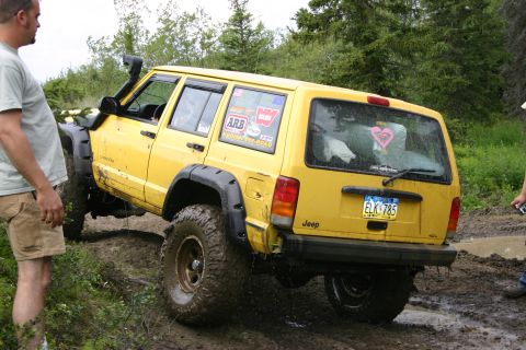 2003 Jeep TJ - A yellow Jeep Cherokee XJ navigates a muddy off-road course, equipped with a snorkel and large tires.