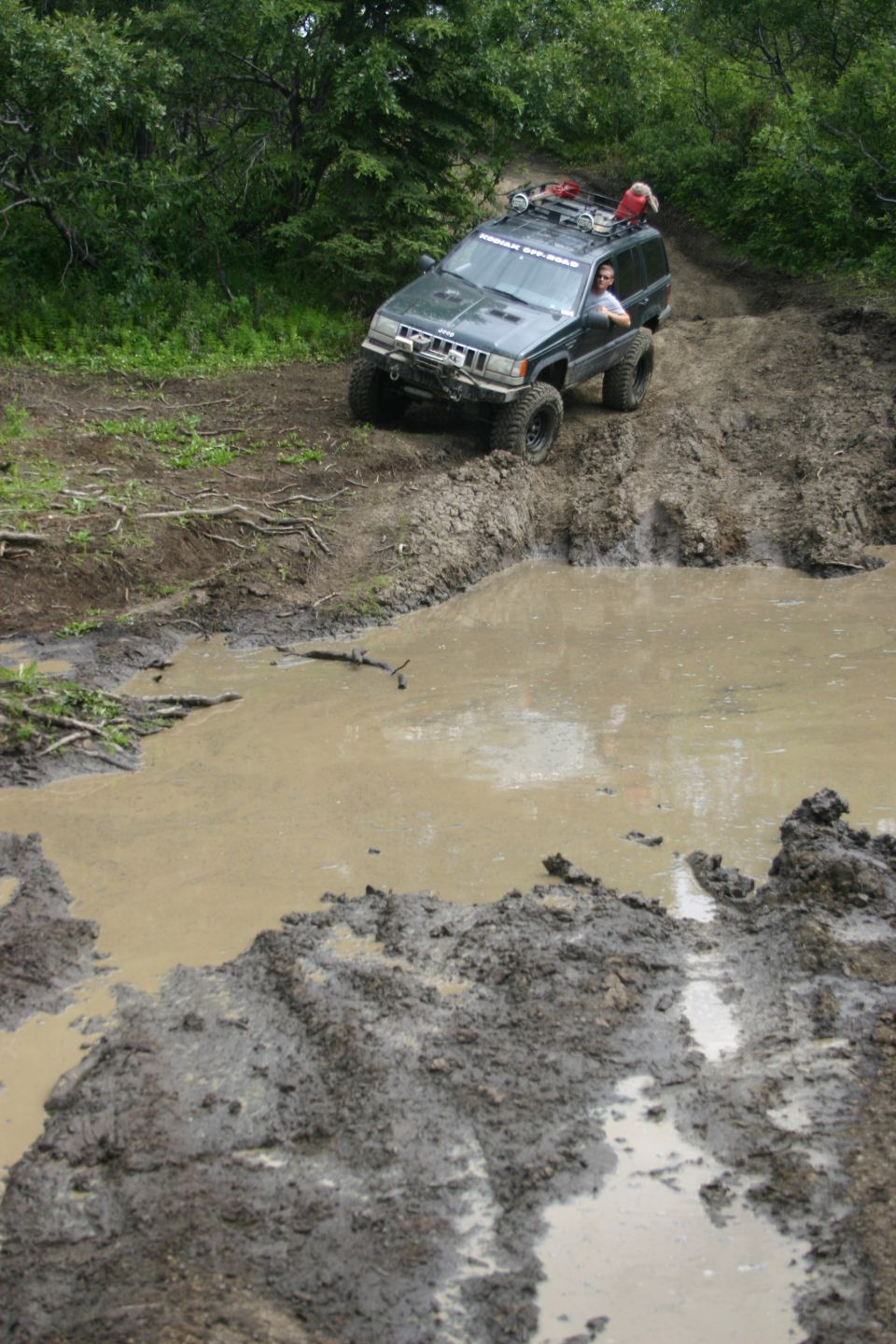 Dark green Jeep Grand Cherokee driving through a muddy trail and large puddle. Driver visible