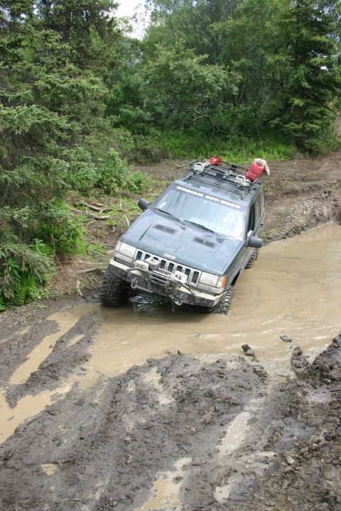 2003 Jeep TJ - Jeep Grand Cherokee navigating a challenging muddy off-road course with a roof rack carrying extra fuel.