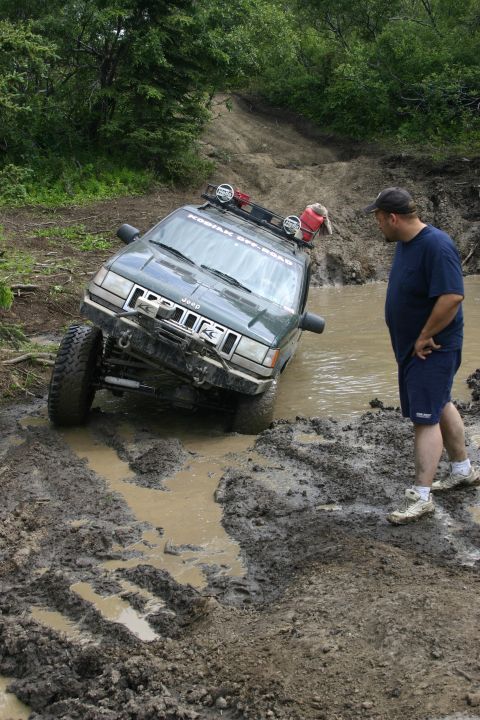 2003 Jeep TJ - A man observes a green Jeep Grand Cherokee partially submerged in a muddy puddle on an off-road trail.