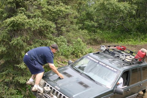 2003 Jeep TJ - A man climbs on the hood of a dark green Jeep Grand Cherokee as it navigates an off-road trail.