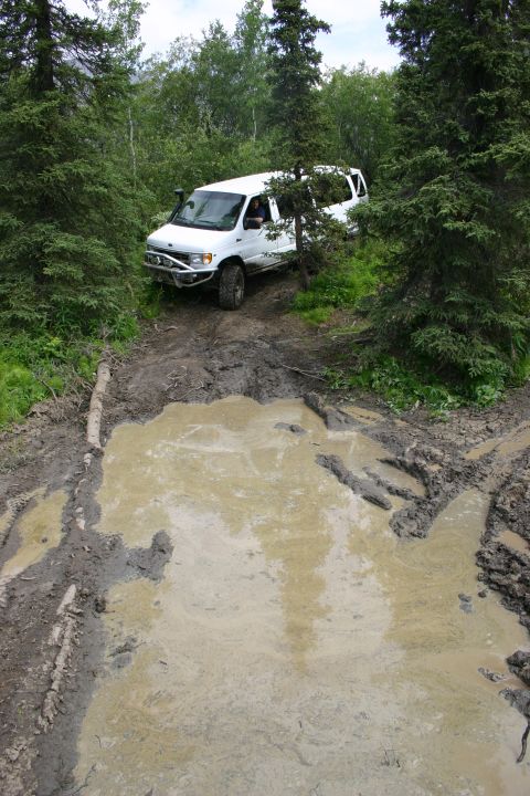 2003 Jeep TJ - A white van navigates a challenging muddy path surrounded by dense trees.