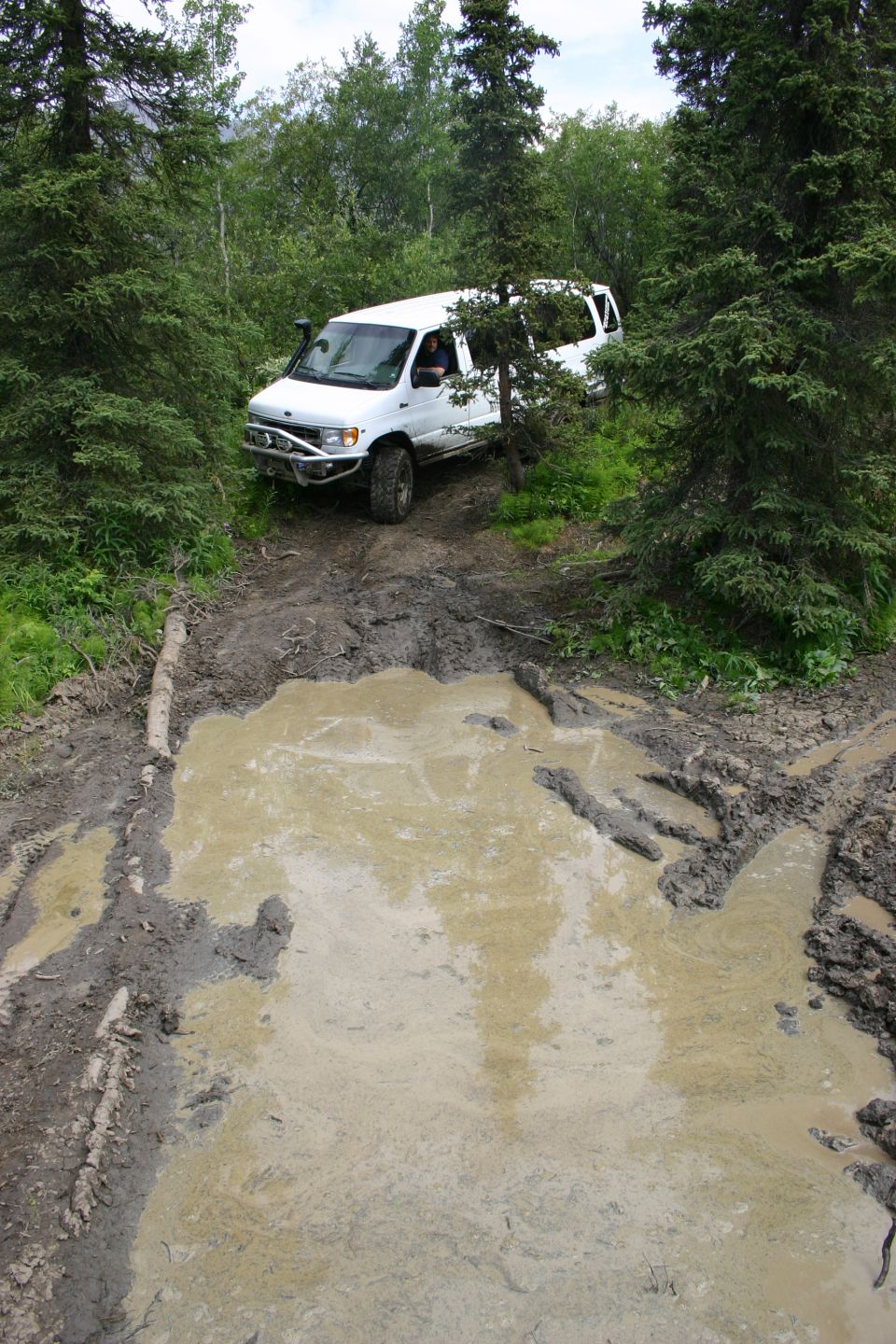White van with off-road tires and bumper driving through a muddy forest trail
