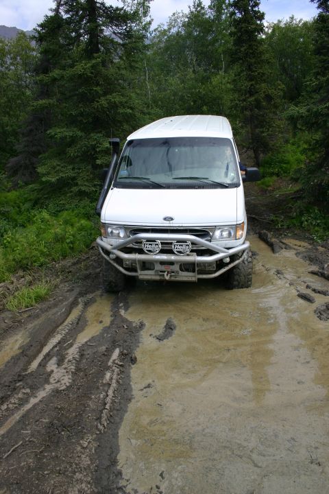 2003 Jeep TJ - A white van navigates a muddy off-road trail, equipped with a snorkel and a heavy-duty bull bar.