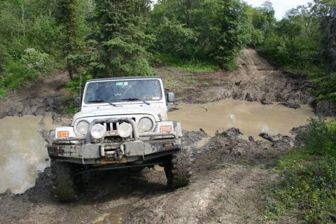 2003 Jeep TJ - A white Jeep TJ navigates a challenging muddy off-road trail, with its front tires submerged in a large puddle.
