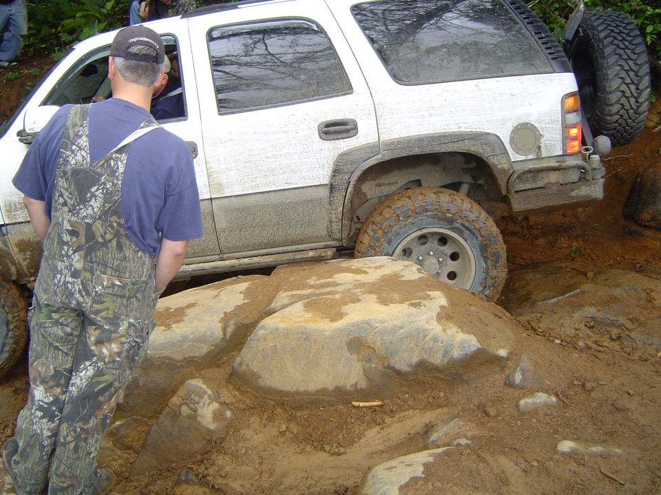 Muddy white Chevy Tahoe crawling over rocks, man in camo bibs watching