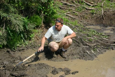 2003 Jeep TJ - A man kneels and uses a shovel to dig in a muddy area surrounded by trees and exposed roots.