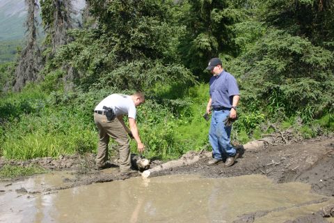 2003 Jeep TJ - Two men are in a muddy, wooded area, one examining something near a puddle while the other stands nearby.