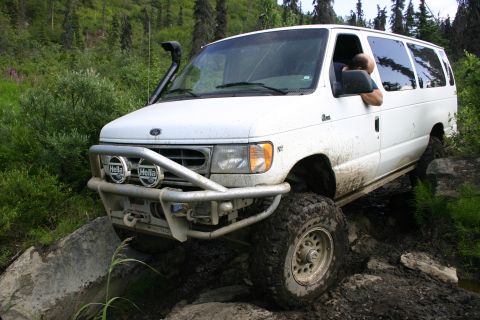 2003 Jeep TJ - A white van equipped for off-roading navigates a challenging rocky and muddy path in a wooded area.