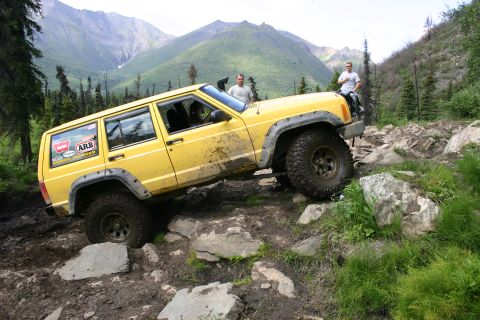 2003 Jeep TJ - A yellow Jeep Cherokee XJ navigates a challenging rocky terrain, with two people visible near the vehicle.