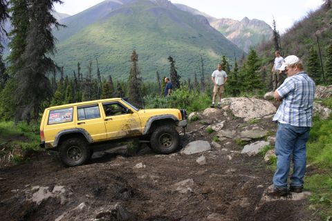 2003 Jeep TJ - A yellow 2003 Jeep Cherokee XJ navigates a challenging muddy off-road course with steep, forested mountains in the background.
