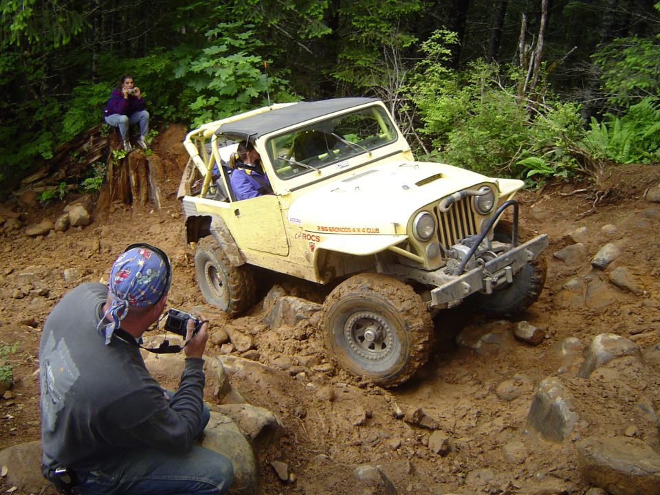 Cream-yellow Jeep with roll cage and winch crawling over rocks on a muddy trail