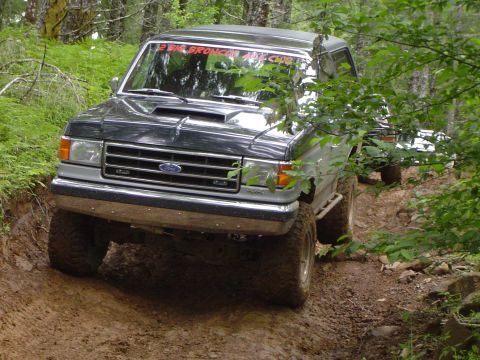 1989 Ford Bronco - A dark gray Ford Bronco navigates a wooded, muddy trail with another vehicle visible behind it.