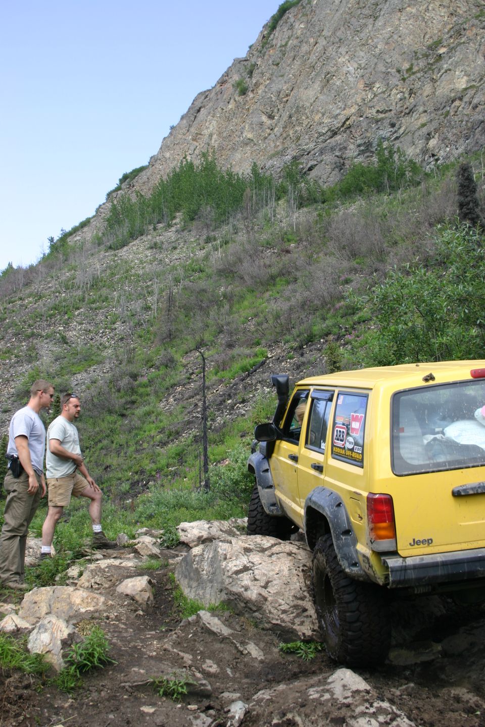 Yellow Jeep Cherokee XJ on rocky trail, two men standing nearby. Jeep has snorkel and stickers on rear window