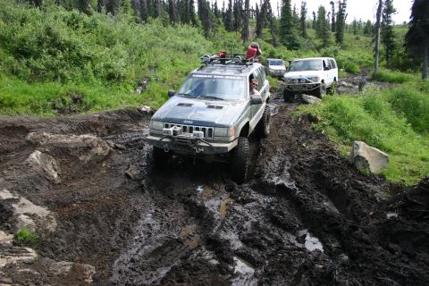2003 Jeep TJ - Two SUVs tackle a challenging, muddy mountain trail surrounded by dense forest.
