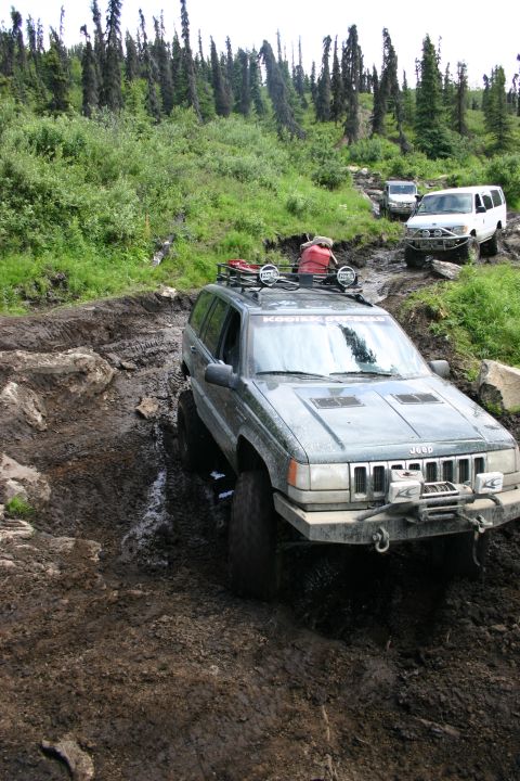 2003 Jeep TJ - Multiple off-road vehicles tackle a challenging, muddy mountain path surrounded by dense forest.