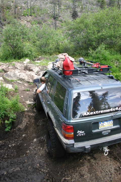 2003 Jeep TJ - A Jeep Grand Cherokee navigates a challenging muddy off-road trail with a red gas can secured to its roof rack.