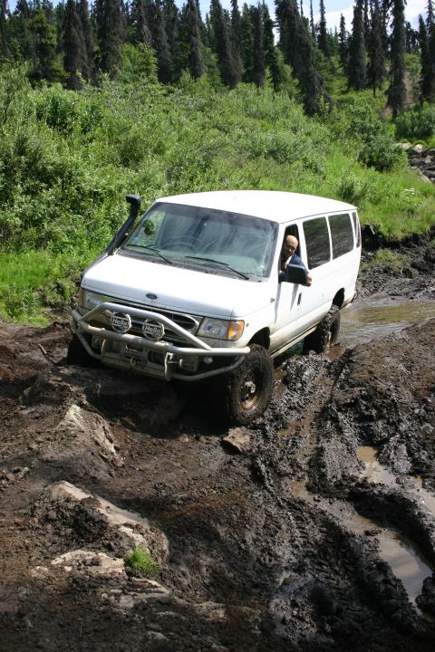 2003 Jeep TJ - A white van navigates a challenging muddy terrain with trees in the background.