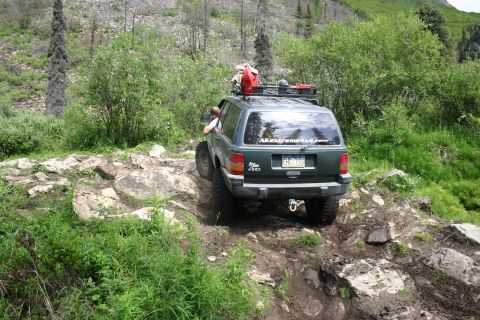 2003 Jeep TJ - A green Jeep Grand Cherokee navigates a challenging, rocky off-road path, equipped with a roof rack and a spare gas can.