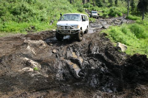 2003 Jeep TJ - Two vehicles struggle on a challenging, muddy off-road path with dense green foliage.