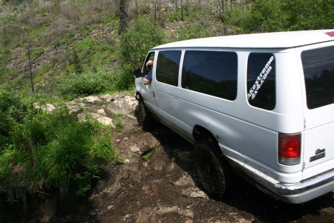 2003 Jeep TJ - A white van with an off-road sticker tackles a challenging, muddy, and rocky terrain.