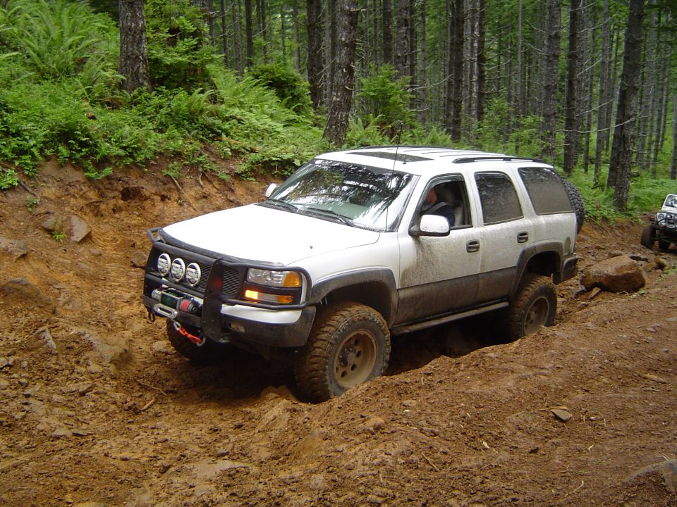 Lifted white Chevy Tahoe with winch bumper and off-road lights on muddy forest trail