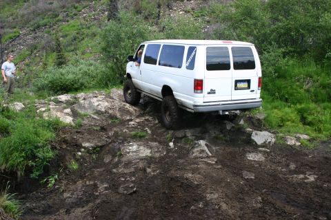 2003 Jeep TJ - White van navigates a challenging rocky and muddy off-road path with a person observing nearby.