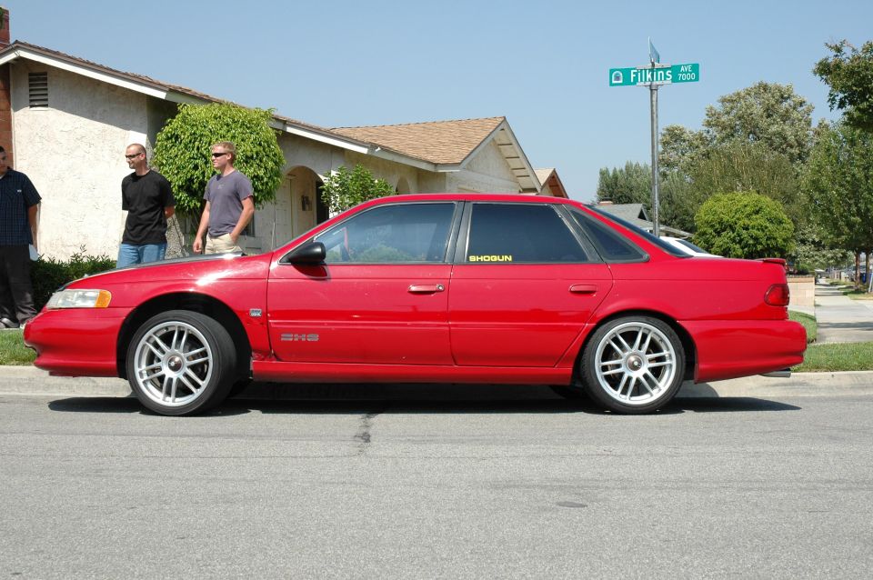 Side view of a red sedan with custom wheels and a 'SHOGUN' sticker on the rear window. Three men stand near a house in the background