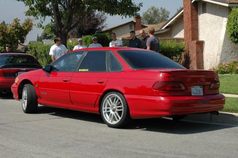 1993 Ford Taurus - A red sedan is parked on a residential street with several people standing nearby.