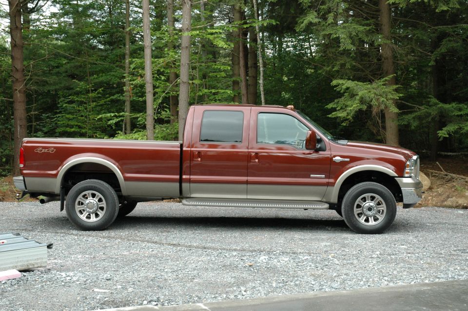 Maroon and tan Ford crew cab long-bed diesel pickup, side view on gravel by woods